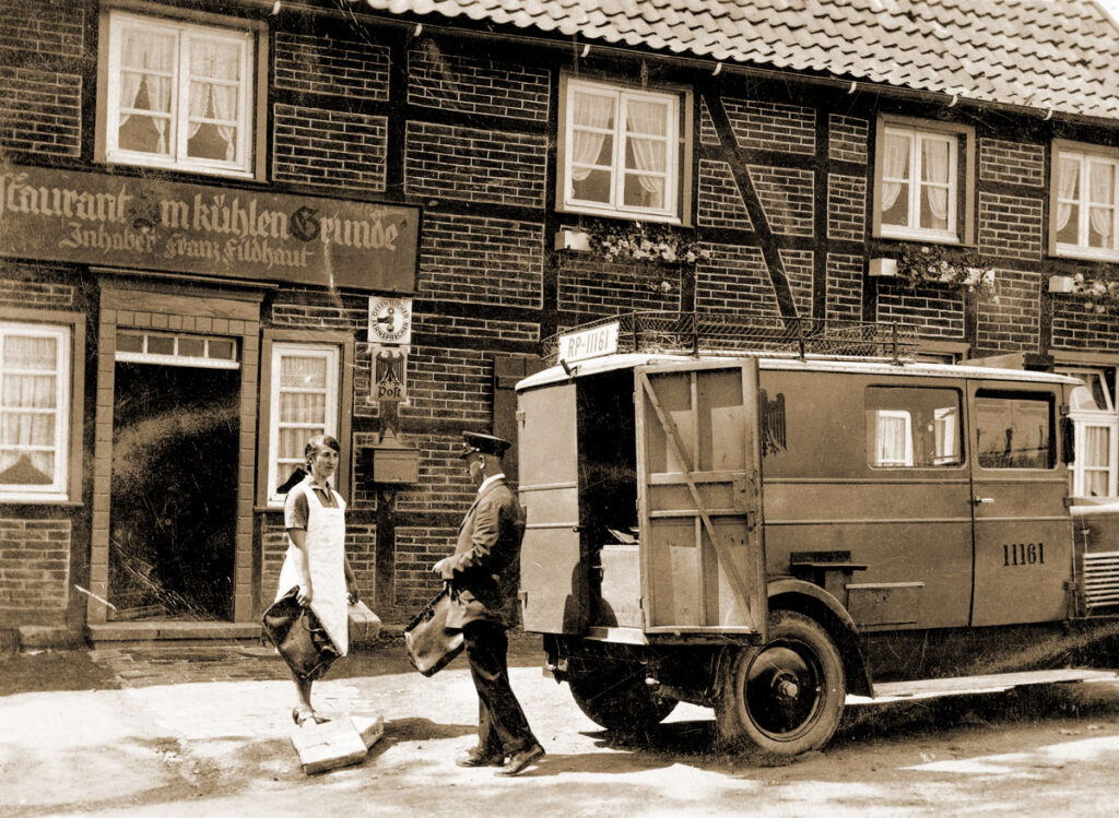 Vor einem Restaurnt steht ein Postwagen. Eine junge Frau erhält von dem Post-Bediensteten etwas.