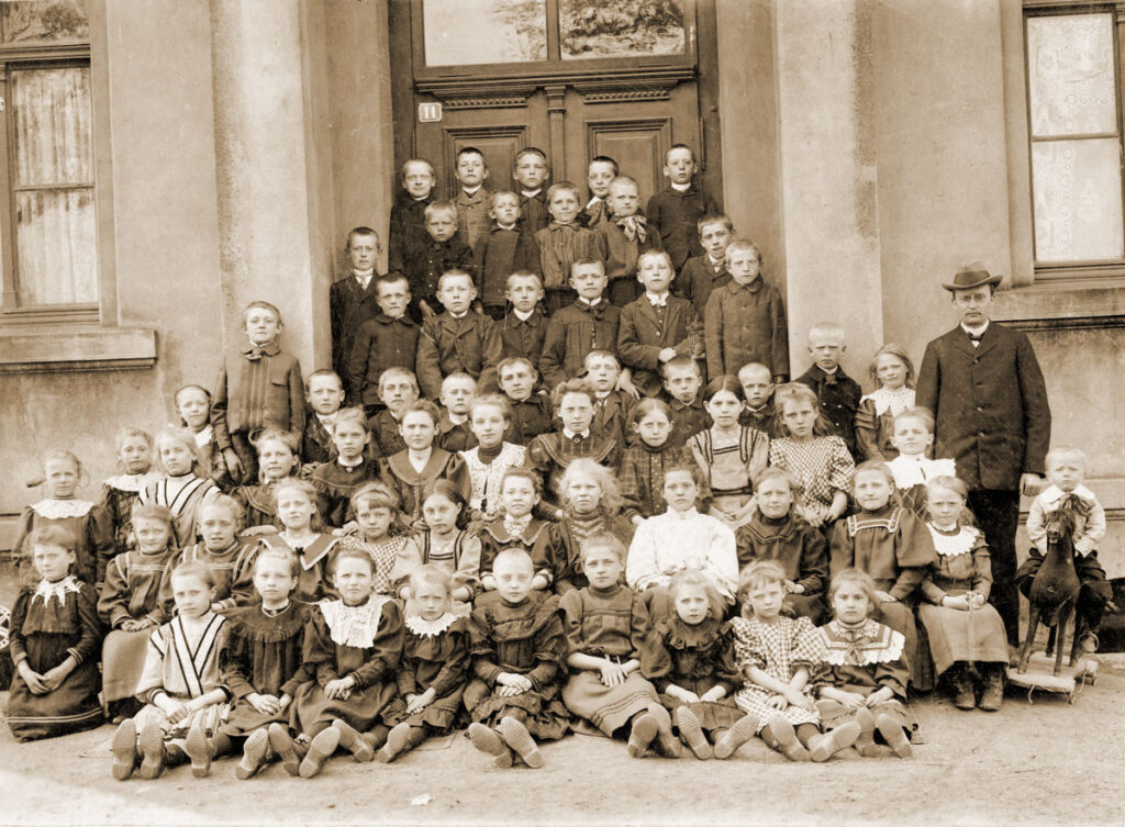 Eine Gruppe Kinder vor einem Gebäude, rechts ihr Lehrer mit Hut