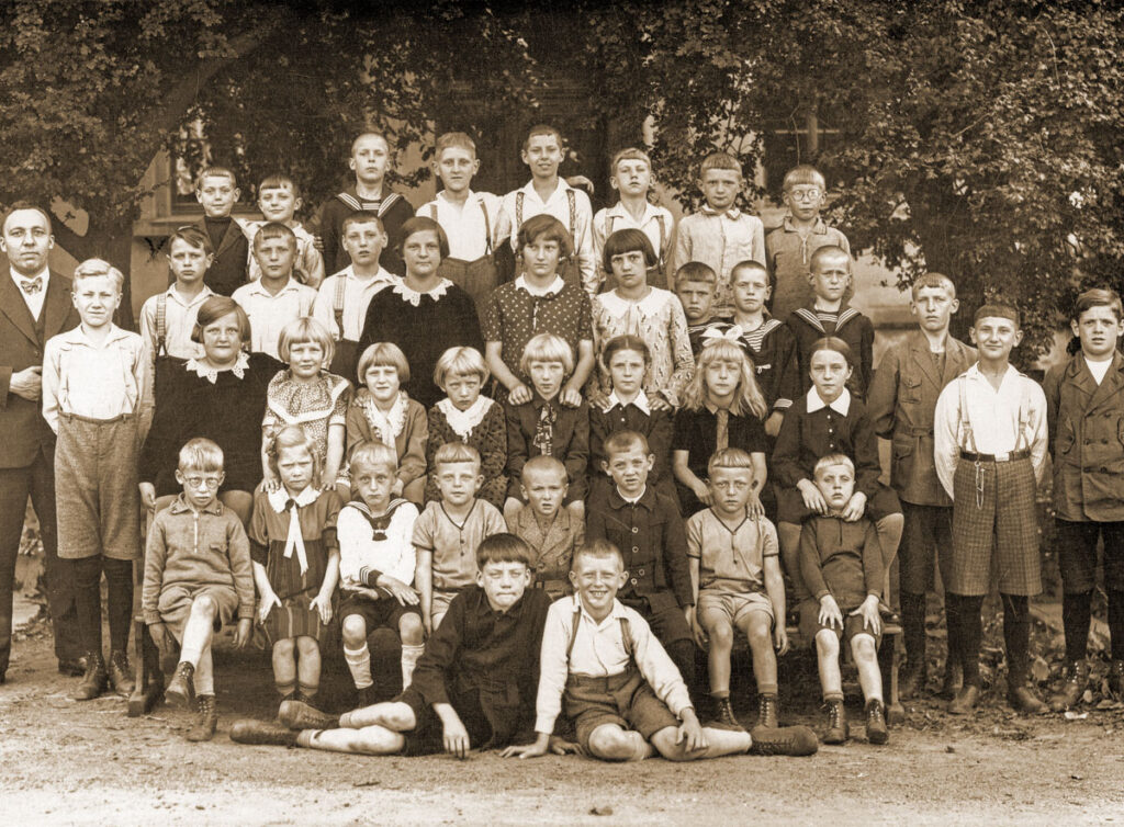 Eine Gruppe Schulkinder steht unter mächtigenb Bäumen, links ihr Lehrer